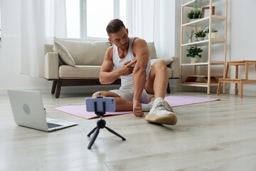 Man sports, watching tape of workout on phone and repeating exercises sports blogger with laptop training online, pumped up man fitness trainer works out at home, the concept of health and body beauty