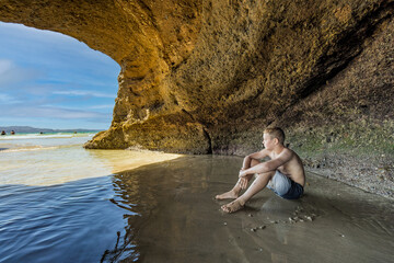 Nature and Wonder: An HDR Photo of a Beautiful Young Boy Sitting in a Cave on the Beach Admiring the Ocean