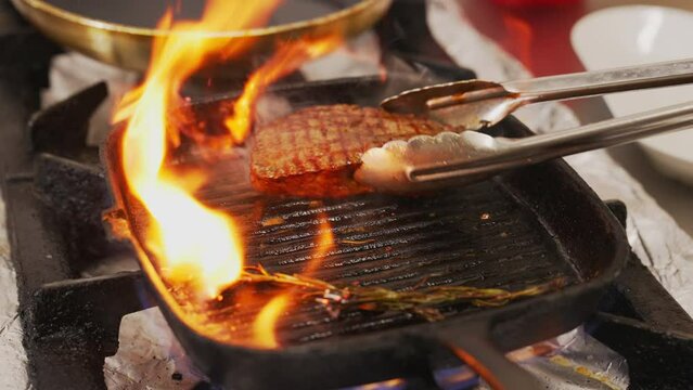 Frying process of striploin steak. Piece of medium rare piece of meat on grill frying pan in flames being turned over with metal cooking tongs. High quality 4k footage