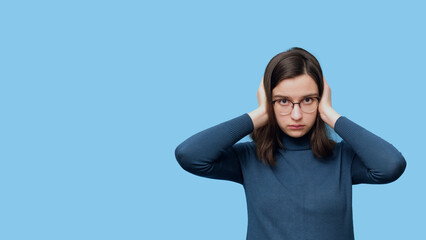 Portrait of a serious student girl in glasses and a sweater covering her ears with her hands in isolation on a blue