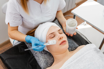 Professional beautician in salon provides comprehensive facial skin care for young woman. Hands of cosmetology specialist applying gold facial mask using brush, making skin hydrated and face glowing.