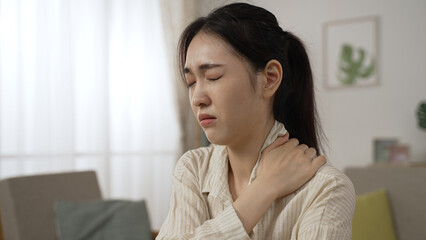 asian female freelance worker having a sudden neck crick is massaging her stiff shoulders and relaxing her muscles while working at home in the living room