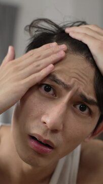 Vertical Screen: upset Asian man facing camera like mirror is worried about his hair loss problem while arranging hairstyle and getting ready in the bedroom at home.