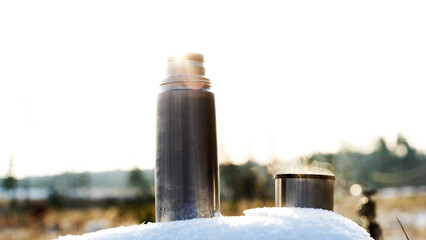 Metal thermos with a hot drink on the background of a winter mountain waterfall. Insulated drink container in the snow