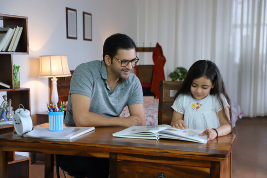 Portrait of smiling father helping cute daughter reading a storybook - Childhood  Parenting Concept. Royalty free image of a happy father sitting with a little beautiful daughter on chair - helping...