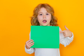 Fototapeta na wymiar Funny kid showing index finger on green empty sheet of paper, isolated on yellow background. Portrait of a kid holding a blank placard, poster. Surprised face, amazed emotions of child.