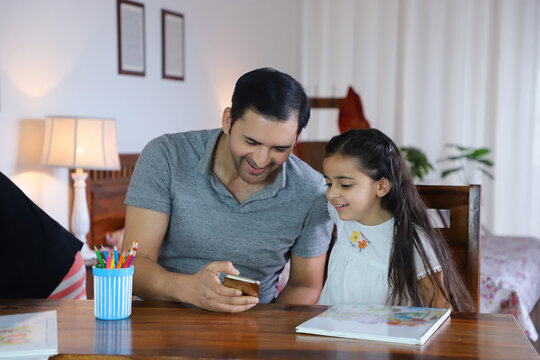 Image of an Indian father and daughter watching a video in mobile phone / smartphone - Parenting  Love  Care. Royalty free image of a pretty daughter and father - wearing casuals and sitting togeth...