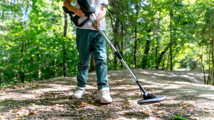 serious boy with a metal detector examines the soil on a rocky trail on a summer evening