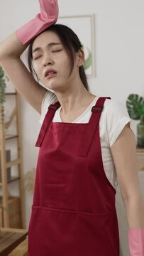 Vertical Screen: overheated asian female wearing household cleaning gloves and apron is wiping sweat on forehead and fanning with hand while vacuuming in the living room