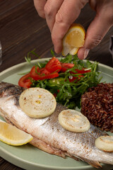 Baked sea bass with brown rice and fresh salad, a man's hand squeezes a slice of lemon