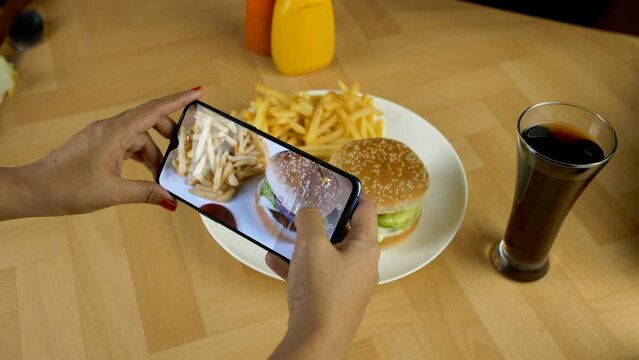 Female clicking pictures of a burger and french fries kept on a plate - social media posts  posting a picture  image sharing. Burger and salty potato fries served with a glass of cold drinks - aera...