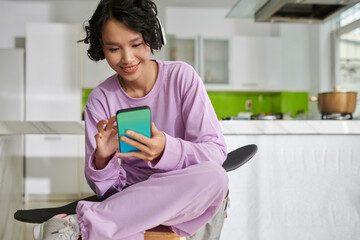 Joyful teenage with skateboard texting friends when sitting on stool in kitchen © DragonImages