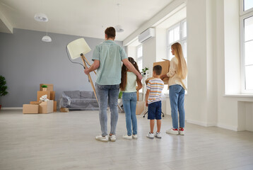 Happy young family with two children moves into their new spacious apartment house. Rear view of family of four standing with cardboard boxes and belongings and inspecting new living room.