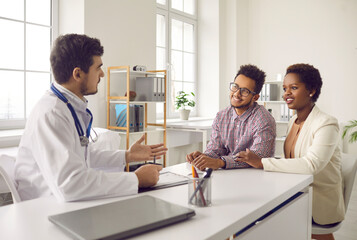 Male gynecologist talk consult multiethnic couple at meeting in private clinic. Smiling man doctor speak with multiracial patients at consultation in hospital. Good medicine, healthcare concept. - obrazy, fototapety, plakaty