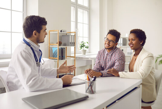 Male gynecologist talk consult multiethnic couple at meeting in private clinic. Smiling man doctor speak with multiracial patients at consultation in hospital. Good medicine, healthcare concept.