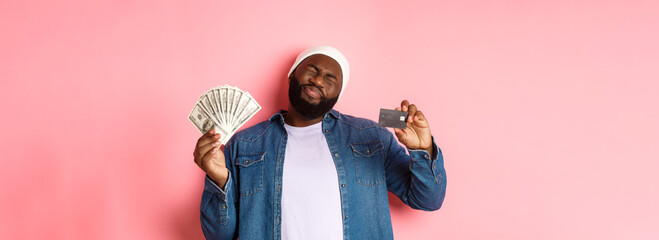 Shopping concept. Sad and whining Black guy showing credit card and dollars, grimacing reluctant, standing over pink background