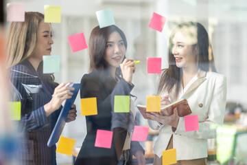 Young creative business Asian woman standing with her colleagues writing new ideas on sticky notes over glass wall, business marketing strategy or User experience ux ui concept.