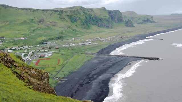 Static landscape view of the small town of Vík í Mýrdal, Iceland during a cloudy day, with grassfields and black sand beach.