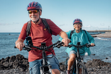 Carefree caucasian senior couple wearing helmets riding on the beach with electric bicycles....