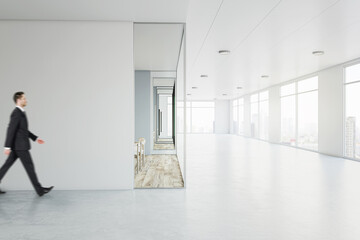 Handsome businessman walking by white wall in sunlit spacious office hall with lots of windows with...