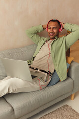 Headphones Man Listening Music. Cheerful Guy Using Laptop At Home. Smiling Millennial Man Sitting On Couch In Living Room And Using Pc Computer, Listening To Music In Wireless Headset 