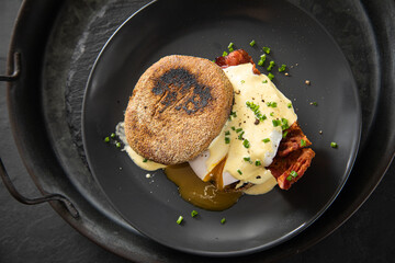 Eggs Benedict mit pochierten Eiern, Sauce Hollandaise, Bacon, Schnittlauch, English Muffin und Cheddar Käse zum Frühstück oder Brunch auf Teller schwarz, Metall Tablet und dunkel Schiefer Hintergrund