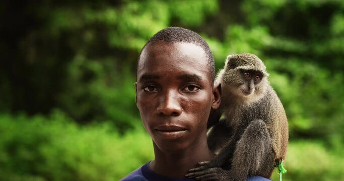 Close Up Portrait of an Authentic African Man Looking at the Camera and Carrying a Little Monkey with Greenery in the Background. Black Male Rural Environment Activist Sending a Call to Save Nature 