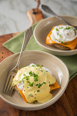Eggs Benedict vegetarisch mit pochierten Eiern, Sauce Hollandaise, Schnittlauch, English Muffin und Cheddar Käse zum Frühstück oder Brunch auf Teller hell mit Serviette grün, Silber Gabel, Holzbrett u