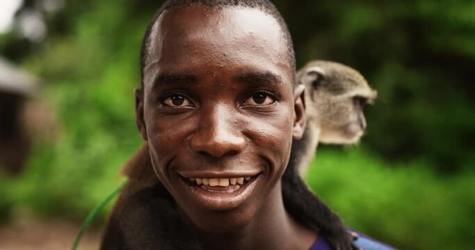 Close Up Portrait of an Authentic African Man Looking at the Camera and Carrying a Little Monkey with Greenery in the Background. Black Friendly Male Smiling and Enjoying the Nature in his Village
