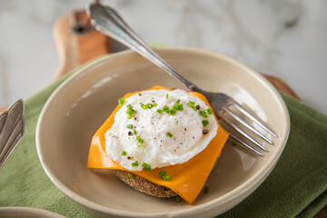 Pochiertes Ei auf English Muffin und Cheddar Käse mit Schnittlauch zum Frühstück oder Brunch auf Teller hell mit Serviette grün, Silber Gabel, Holzbrett und Marmor Hintergrund