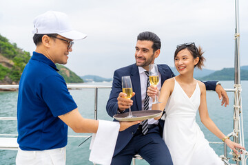 Asian man waiter serving champagne to passenger tourist while private catamaran boat yacht sailing...