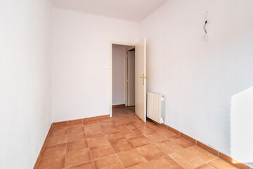 An empty spacious room with an open door, white walls and ceiling, caramel-colored square floor tiles and a radiator on the wall for warmth in the cold season. The room is lit by bright daylight.