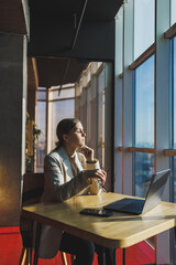 Young content worker in casual clothes sitting at a desk and using a netbook while working on the background of a spacious office. Business woman in the office works sitting near the window