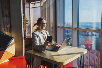 Cheerful young woman in casual wear having telephone conversation sitting at working place near laptop in office, smiling female freelancer calling to colleague for share good news about business