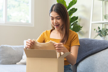 Happy excited, asian young woman, girl customer sitting on sofa at home, open and unpack cardboard box carton parcel after buy fashion clothes, ordering present and shopping online, delivery service.