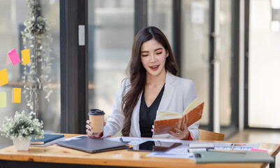 Asian Businesswoman having a tea, sitting on desk in workplace, writing down notes, opened laptop in document the financial report, business plan investment, finance analysis concept.
