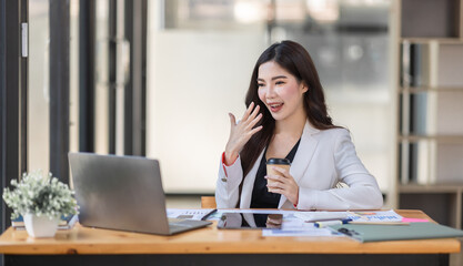 Fototapeta na wymiar Asian Businesswoman having a tea, sitting on desk in workplace, writing down notes, opened laptop in document the financial report, business plan investment, finance analysis concept.