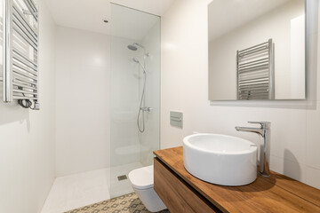 Fototapeta na wymiar Bathroom with small round white sink on long wooden countertop floating in air. Mirror above round sink reflects radiator heater on opposite wall. Shower area is separated by glass railing.