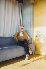 Young male worker in casual clothes and glasses looking at laptop while sitting on sofa with laptop in bright modern workspace. A successful freelancer works remotely online in a workspace