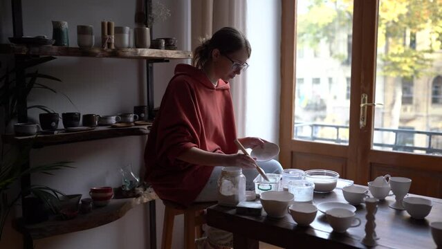 Concentrated introverted woman working alone in pottery making original handmade crockery for sale at crafts fair. Carried away girl in casual clothes sits at table applies white paint to plates