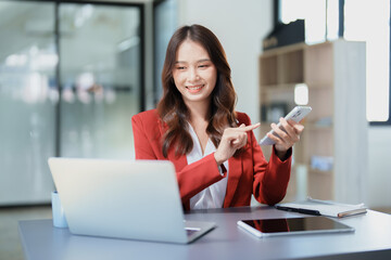 Portrait of a young Asian woman showing a smiling face as she uses her phone, computer and financial documents on her desk in the early morning hours
