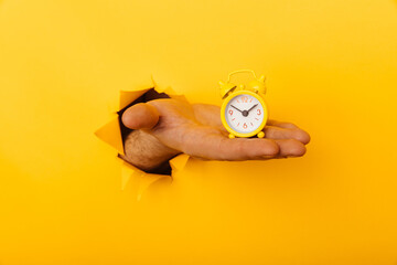 Hand with mini alarm clock through a paper hole in yellow background close-up