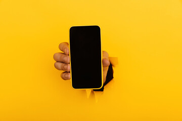 Hand with a smartphone out of a hole torn in yellow paper wall. Advertising concept