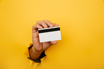 Male hand is holding credit bank card in torn hole of yellow background. Online shopping, purchasing products concept.