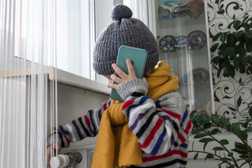 A small child warmly dressed in a sweater and a hat turns the tap on the radiator. The concept of...