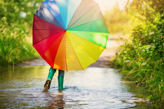 Boy walking through the puddle with colorful umbrella in his hands.