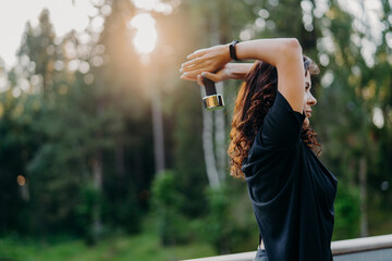 Profile shot of active motivated young woman dressed in black t shirt, raises dumbbell over head,...