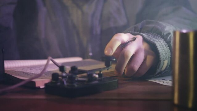 Man in jacket writes down information in notebook. Male person sends signals in Morse code by telegraph sitting at table near mug close view
