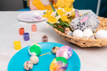 Fototapeta na wymiar Close up of wicker basket with daffodils and plates of painting eggs on the table. Preparation for the celebration of Easter, eggs dyeing