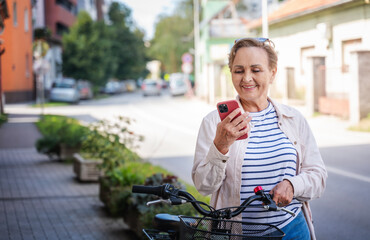 Beautiful happy caucasian active senior woman 60 years with bicycle in summer city using smartphone